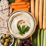 Top view of hummus in bowl with arranged cut vegetables slices, breadsticks, olives and pita bread