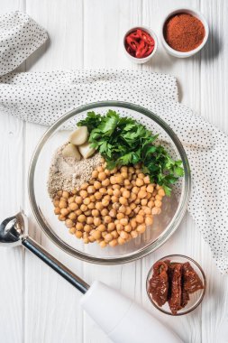 Flat lay with chickpeas and other ingredients for hummus in bowl, blender and dried tomatoes on wooden tabletop clipart