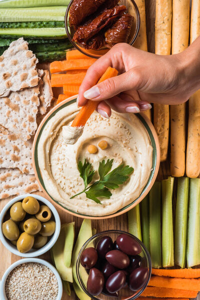partial view of woman and arranged hummus in bowl, pita bread, cut vegetables, dried tomatoes and olives