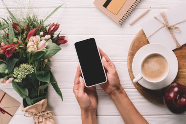 cropped shot of woman holding smartphone with blank screen at surface with wrapped bouquet of flowers and cup of coffee