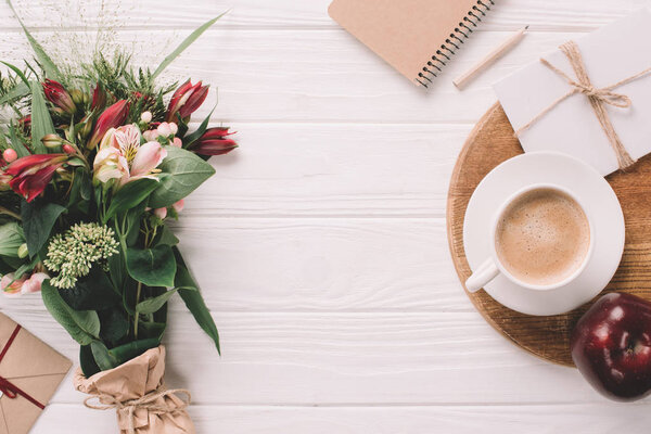 flat lay with wrapped bouquet of flowers, cup of coffee and notebook on white wooden tabletop
