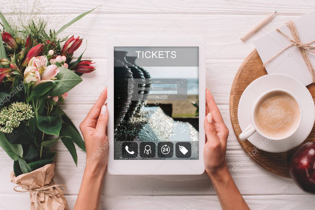 partial view of woman holding tablet with tickets website on screen at surface with cup of coffee and bouquet of flowers