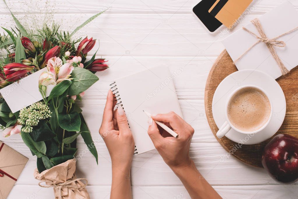 partial view of woman making notes in notebook at tabletop with bouquet of flowers and cup of coffee