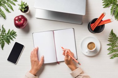 kadın Freelancer dizüstü bilgisayar, akıllı telefon ve eğreltiotu yaprakları ile masada boş ders kitabı yazma kırpılmış görüntü