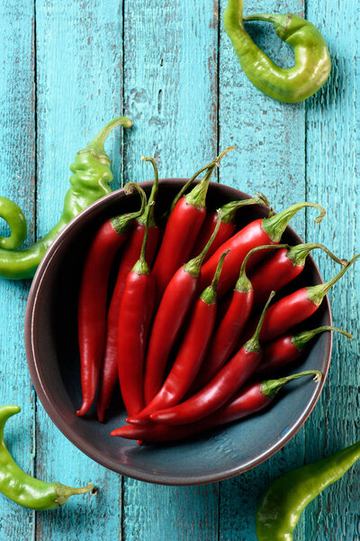 top view of red chili peppers in bowl and scattered green peppers on blue wooden table