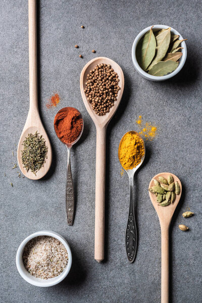 top view of different dried aromatic spices in spoons and bowls on grey  background