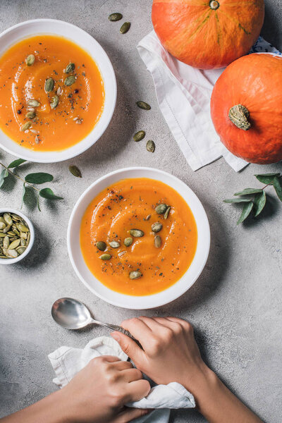 partial view of woman sitting at table with pumpkins and plates with pumpkin soup and seeds 