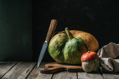close up view of food composition with pumpkins, knife and cutting board arranged on wooden surface on dark background clipart