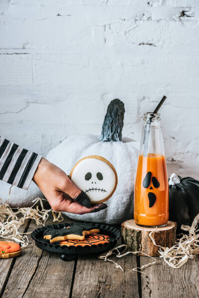 cropped shot of woman holding halloween cookie near pumpkins and juice on wooden surface