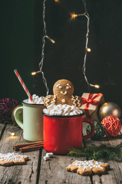 две чашки какао с зефиром и пряничным человечком на деревянном столе с гирляндой на Рождество
