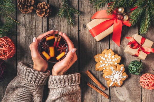 cropped view of woman holding glass with hot mulled wine on wooden background with christmas gifts