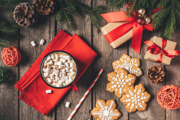 вид сверху на пряники, рождественские подарки и чашку какао с зефиром на деревянном фоне с елкой
