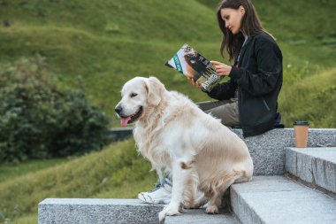 young woman reading magazine while sitting with dog in park clipart