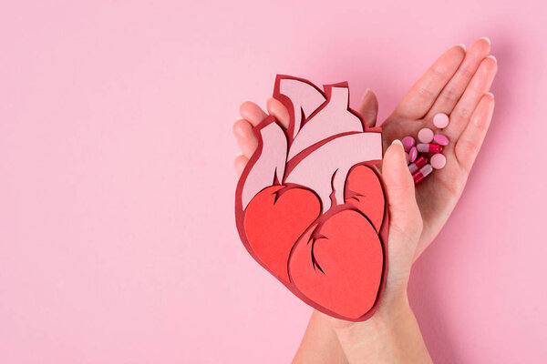 cropped image of woman holding anatomical human heart and various pills on pink 