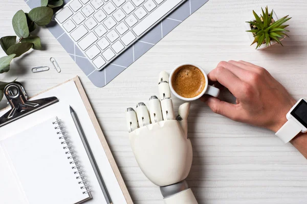 cropped image of businessman with prosthesis hand and smartwatch sitting at table with coffee cup and computer keyboard in office