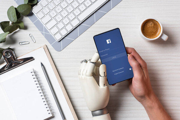 partial view of businessman with prosthesis arm using smartphone with facebook on screen at table with coffee cup in office 