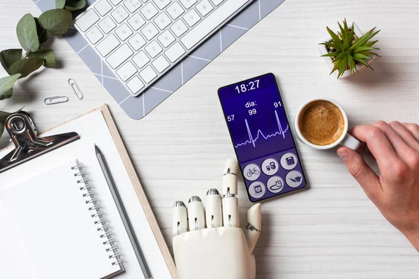 オフィスのコーヒー カップとテーブルで画面上の医療アプリケーションでスマート フォンを使用する人工腕を持ったビジネスマンの部分的なビュー — ストック写真