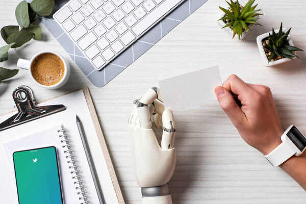 partial view of businessman with cyborg hand and smartwatch holding blank visit card at table with smartphone with youtube on screen in office 