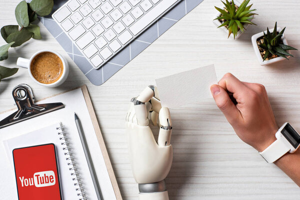 partial view of businessman with cyborg hand and smartwatch holding blank visit card at table with smartphone with youtube on screen in office 