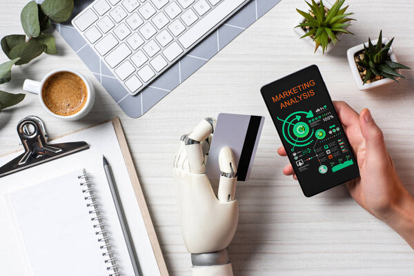 partial view of businessman with prosthesis arm holding credit card and using smartphone with marketing analysis on screen at table in office 