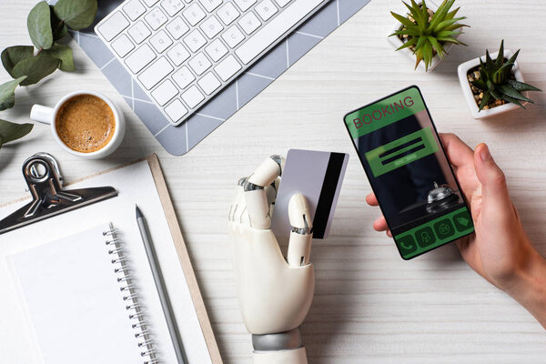 partial view of businessman with prosthesis arm holding credit card and using smartphone with booking on screen at table in office 