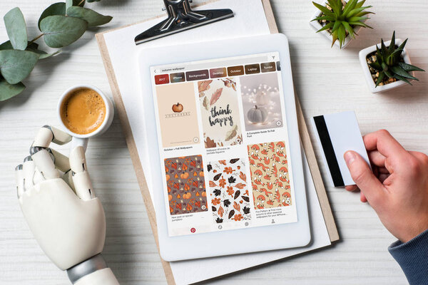 cropped image of businessman with prosthesis hand holding credit card and using digital tablet with pinterest on screen at table with coffee cup in office 