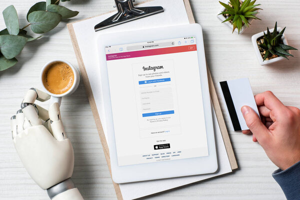 cropped image of businessman with prosthesis hand holding credit card and using digital tablet with instagram on screen at table with coffee cup in office 