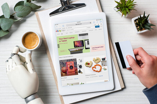 cropped image of businessman with cyborg hand holding credit card and using digital tablet with ebay on screen at table with coffee cup in office 