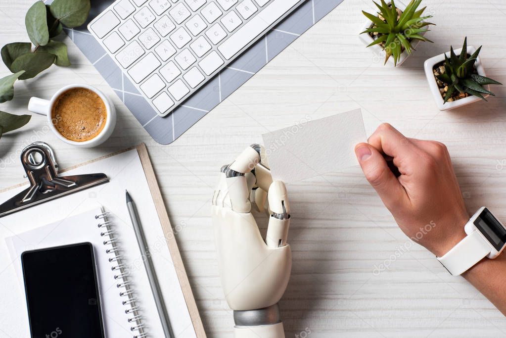 cropped image of businessman with cyborg hand and smartwatch holding blank visit card at table with smartphone with blank screen in office 