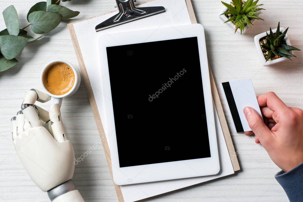 partial view of businessman with prosthesis hand holding credit card and using digital tablet with blank screen at table with coffee cup in office 