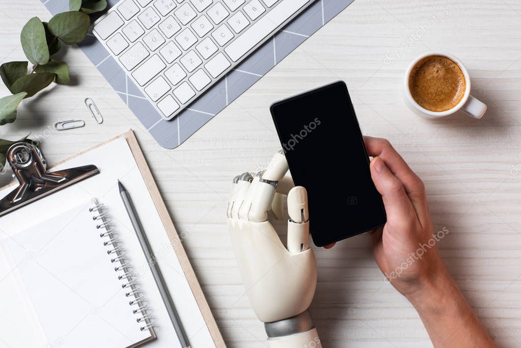 partial view of businessman with cyborg hand using smartphone with blank screen at table with coffee cup in office 
