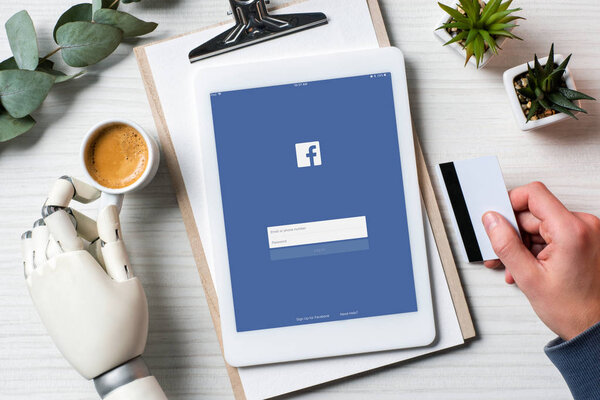 частичный взгляд бизнесмена с протезной рукой, держащей кредитную карту, сидя за столом с чашкой кофе и цифровым планшетом с facebook на экране
