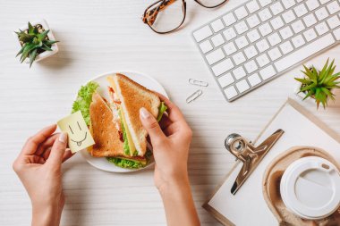 holding kağıt Kupası Office masada sandviç ve kahve ile öğle yemeği gülümseme sembolü işkadını kısmi görünümünü 