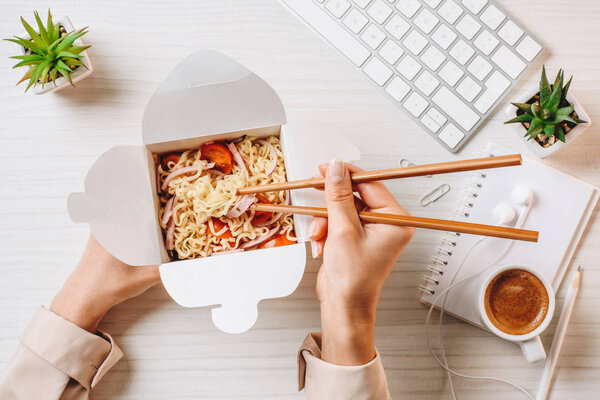 cropped image of businesswoman eating noodle by chopsticks at table with coffee cup and computer keyboard in office 