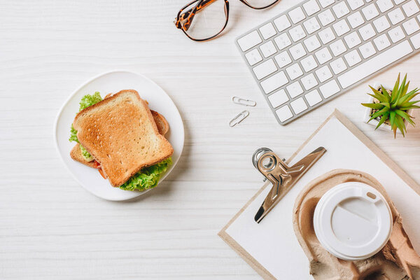 elevated view of paper coffee cup, sandwich and computer keyboard at table in office 