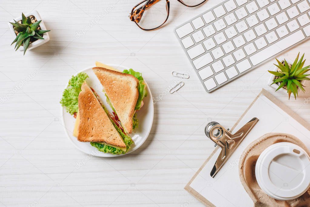 view from above of disposable coffee cup, sandwich, computer keyboard and eyeglasses at table in office 
