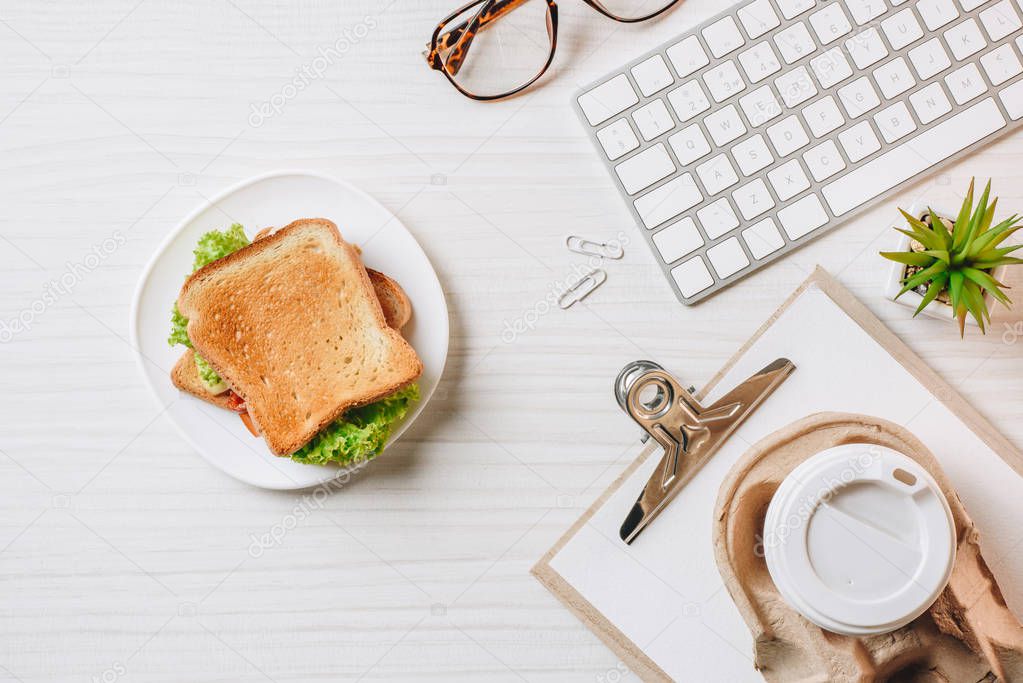 elevated view of paper coffee cup, sandwich and computer keyboard at table in office 