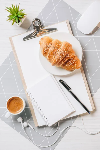 view from above of empty clipboard, earphones, croissant and coffee cup at table in office