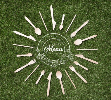 top view of wooden spoons with forks and knives in shape of sun lying on grass, menu inscription clipart