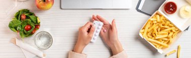 dizüstü bilgisayar, sağlıklı yiyecek ve abur cubur işyerinde hapları tutan kişi kısmi üstten görünüm 