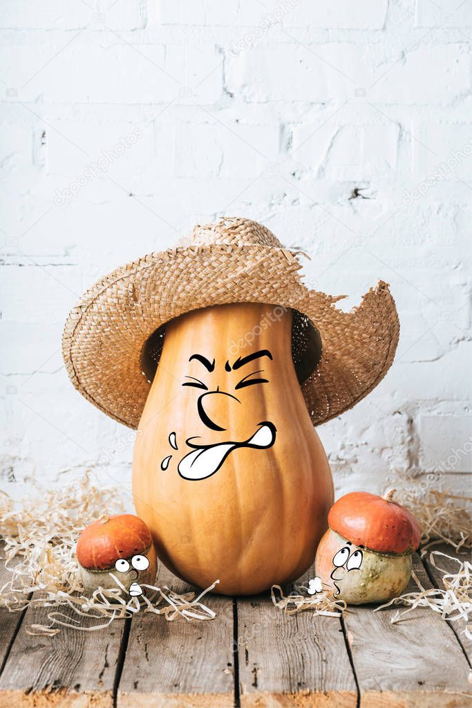 close up view of ripe pumpkins with drawn grimacing facial expression and straw hat on wooden surface and white brick wall backdrop