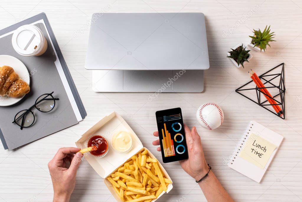 cropped shot of person eating french fries with ketchup and using smartphone with business charts at workplace
