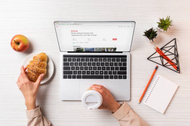 airbnb Web sitesi ile dizüstü bilgisayar kullanırken tek kullanımlık kahve ve kruvasan tutan işkadını kırpılmış atış