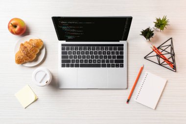 html kodu ile dizüstü ekran, tek kullanımlık kahve fincanı, apple ve kruvasan masada üstten görünüm