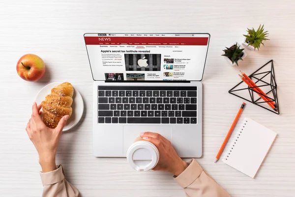 裁剪拍摄的女商人拿着一次性咖啡杯和羊角面包 而使用笔记本电脑与 Bbc 新闻网站 — 图库照片