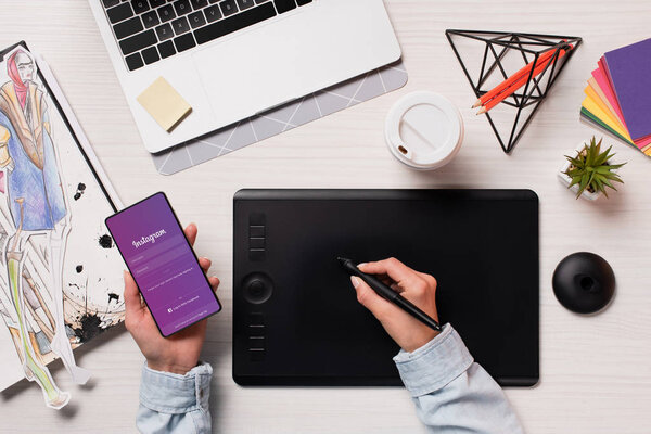 cropped view of designer using graphics tablet, pen and smartphone with instagram app on screen, flat lay