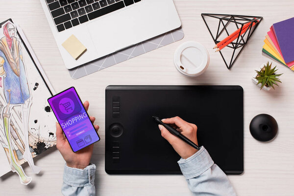 cropped view of designer hands using graphics tablet, pen and smartphone with shopping app on screen, flat lay
