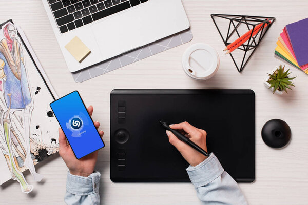 cropped view of designer using graphics tablet, pen and smartphone with shazam app on screen, flat lay