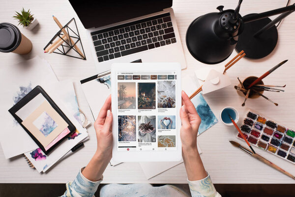 cropped view of designer holding tablet with pinterest website on screen at office desk with art supplies, flat lay
