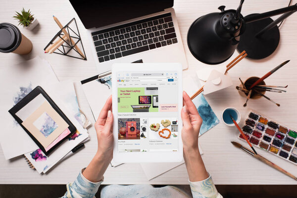 cropped view of designer holding tablet with ebay website on screen at office desk with art supplies, flat lay